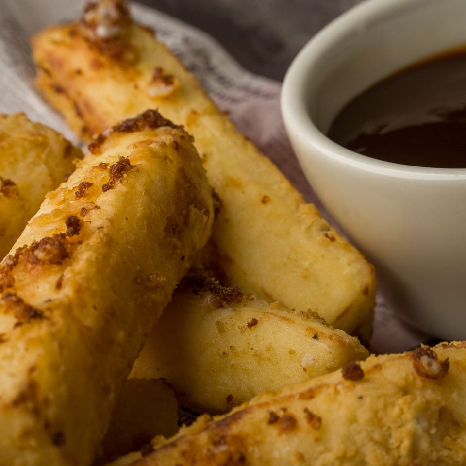 Close-up of golden-brown cheese fries with crispy bits, served with a side of dark dipping sauce.