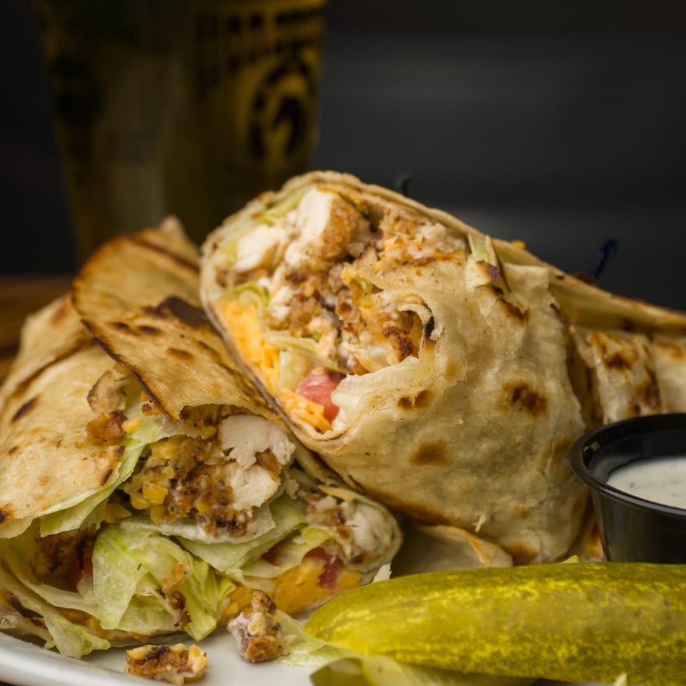 Two crispy chicken wraps with lettuce, cheese, and tomato, served with a pickle and dipping sauce.