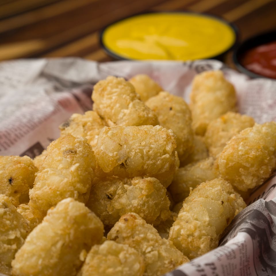 Golden-brown tater tots in a paper-lined basket with mustard and ketchup in the background.