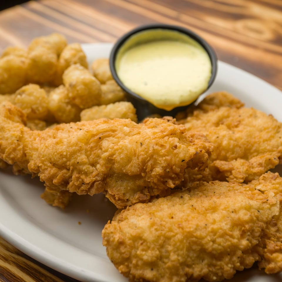 Crispy fried chicken tenders and tater tots with a side of dipping sauce on a white plate.