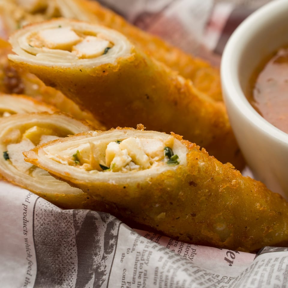 Golden-fried spring rolls with chicken and herbs, served with dipping sauce in a white cup.
