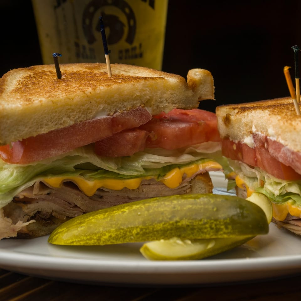 A grilled turkey club sandwich with cheese, lettuce, and tomato, served with a pickle spear.