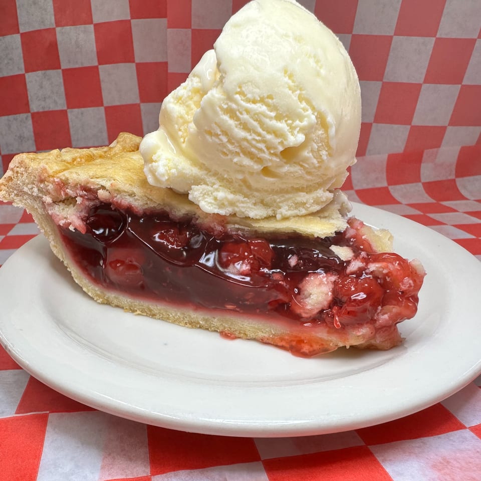 A slice of cherry pie with a scoop of vanilla ice cream on a white plate.