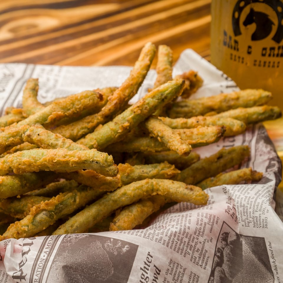 A basket of crispy fried green beans on newspaper, with a Horseshoe Bar & Grill glass in the background.