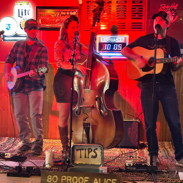A band with a banjo, upright bass, and acoustic guitar performs on a stage with red lighting.