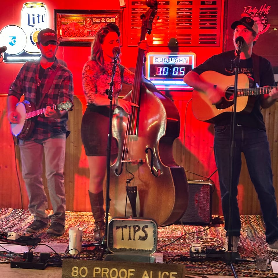 A band with a banjo, upright bass, and acoustic guitar performs on a stage with red lighting.