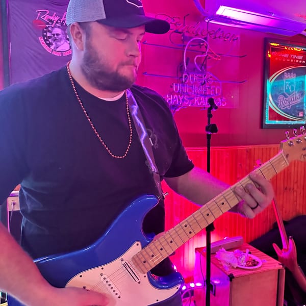 A man in a baseball cap and beaded necklace plays a blue electric guitar under neon lights.