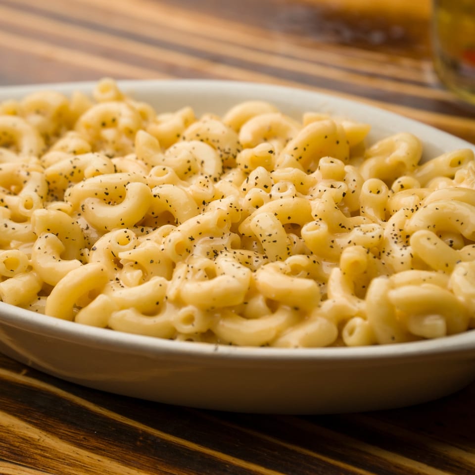 Creamy macaroni and cheese, generously seasoned with black pepper, served in a white bowl on a wooden table.