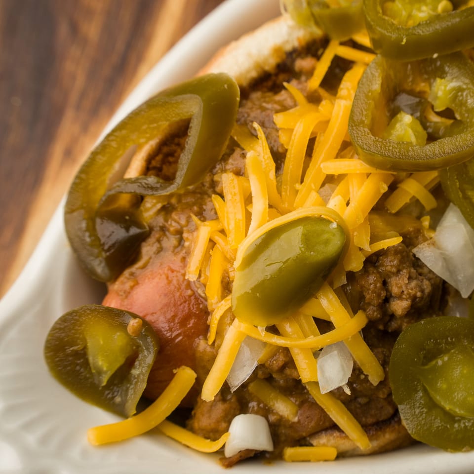Close-up of a chili cheese hot dog with shredded cheddar, diced onions, and sliced jalapeños.