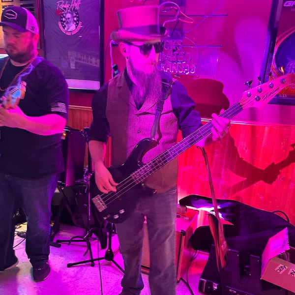 Two musicians performing on stage, one playing bass guitar, under purple and red lighting.