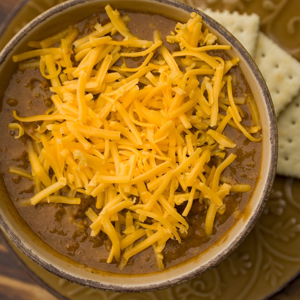 A bowl of chili topped with shredded cheddar cheese, with crackers on the side.