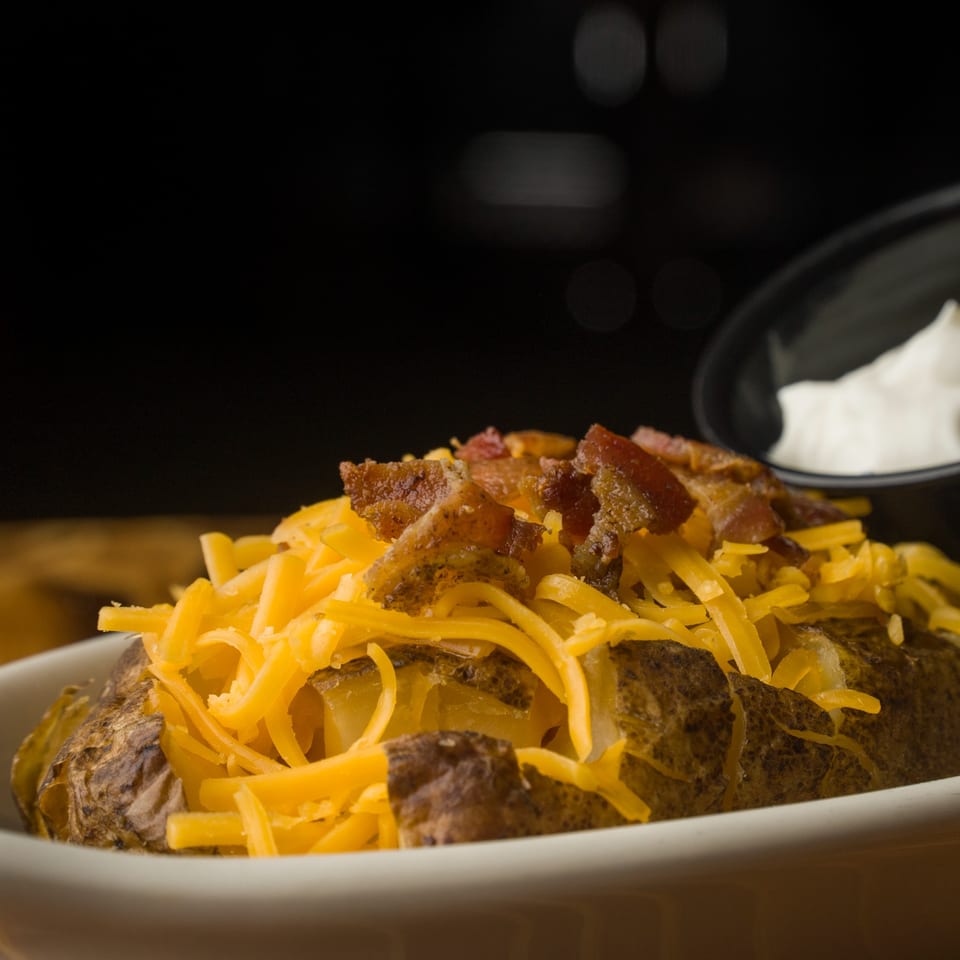 A baked potato topped with shredded cheddar cheese and crispy bacon bits, with sour cream in the background.