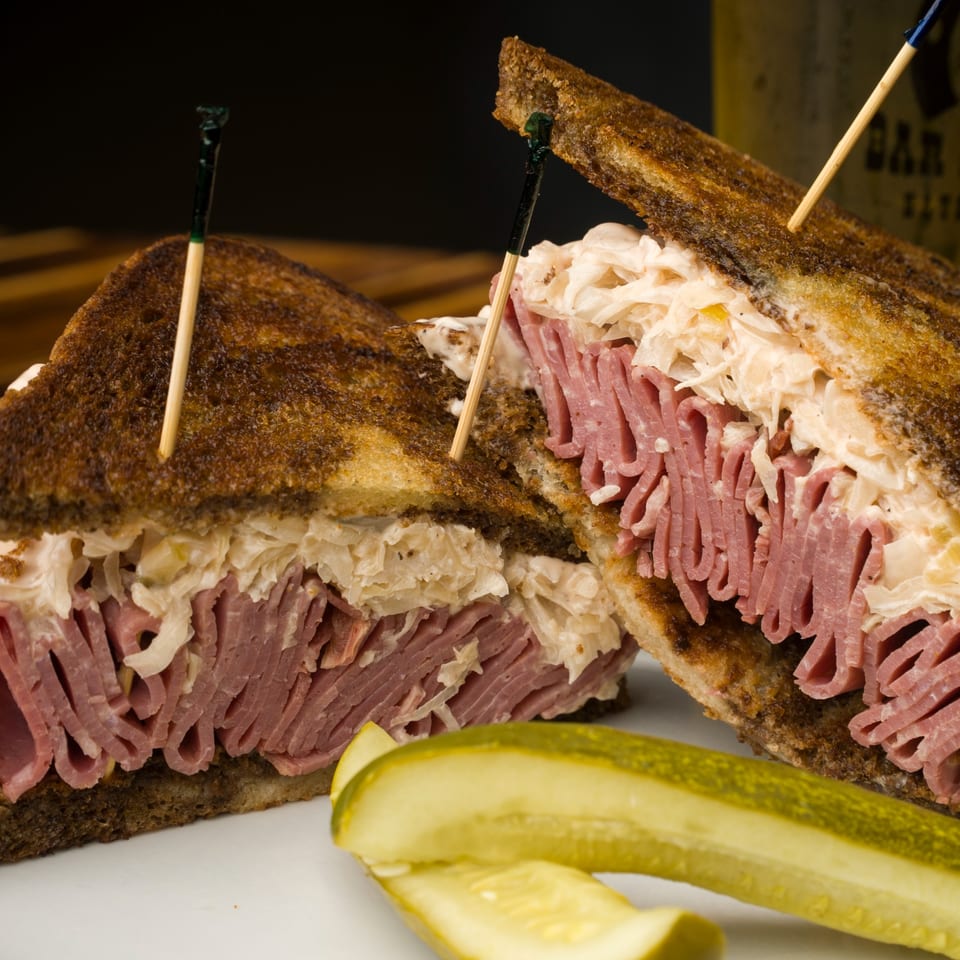 A Reuben sandwich, cut in half, with corned beef, sauerkraut, and pickles on a white plate.