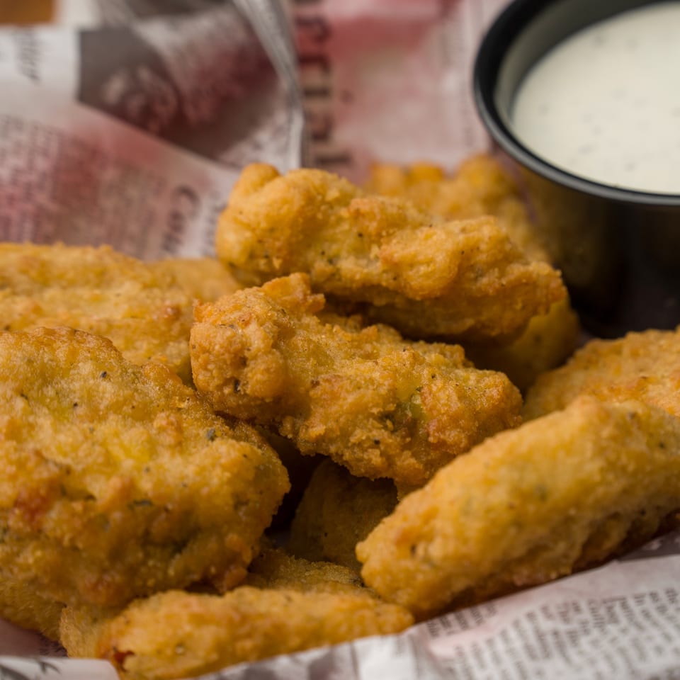 Close-up of golden-brown fried pickles in a basket lined with newspaper, with a side of ranch.